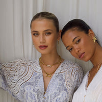 Two women pose calmly before a white curtain, both wearing light patterned tops and elegant gold jewelry from The Songbird Collection, including the Icon Link Necklace. Their hair is pulled back as they look at the camera.