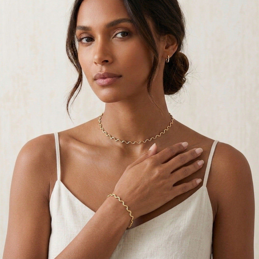 Woman wearing gold jewelry including a necklace, bracelet, and earrings on a neutral background - stainless steel silver wavy chain necklace a marble surface - Aura_Ripple_Chain_Necklace_-_The_Songbird_Collection 