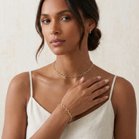 Woman wearing gold jewelry including a necklace, bracelet, and earrings on a neutral background - stainless steel silver wavy chain necklace a marble surface - Aura_Ripple_Chain_Necklace_-_The_Songbird_Collection 