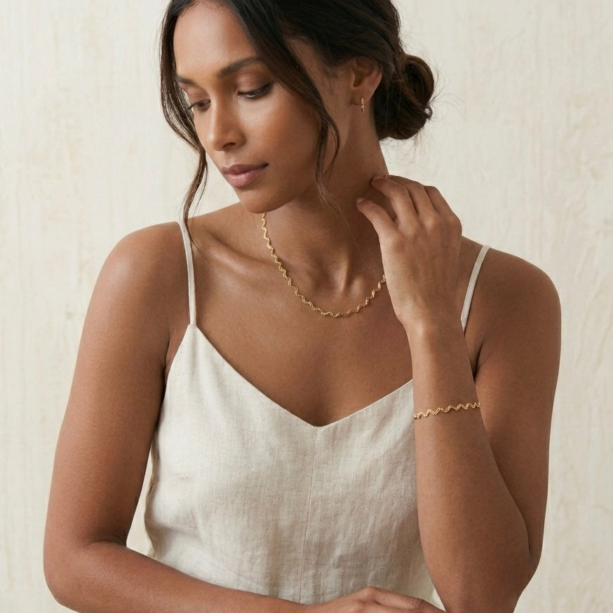 Woman wearing gold jewelry including a necklace and bracelet against a neutral background - stainless steel silver wavy chain necklace a marble surface - Aura_Ripple_Chain_Necklace_-_The_Songbird_Collection 