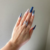 A hand showcasing a silver-colored chandelier ring with cubic zirconia on a finger, with multi-colored nails.