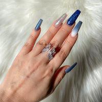 A hand showcasing a silver-colored chandelier ring with cubic zirconia on a finger, with multi-colored nails.