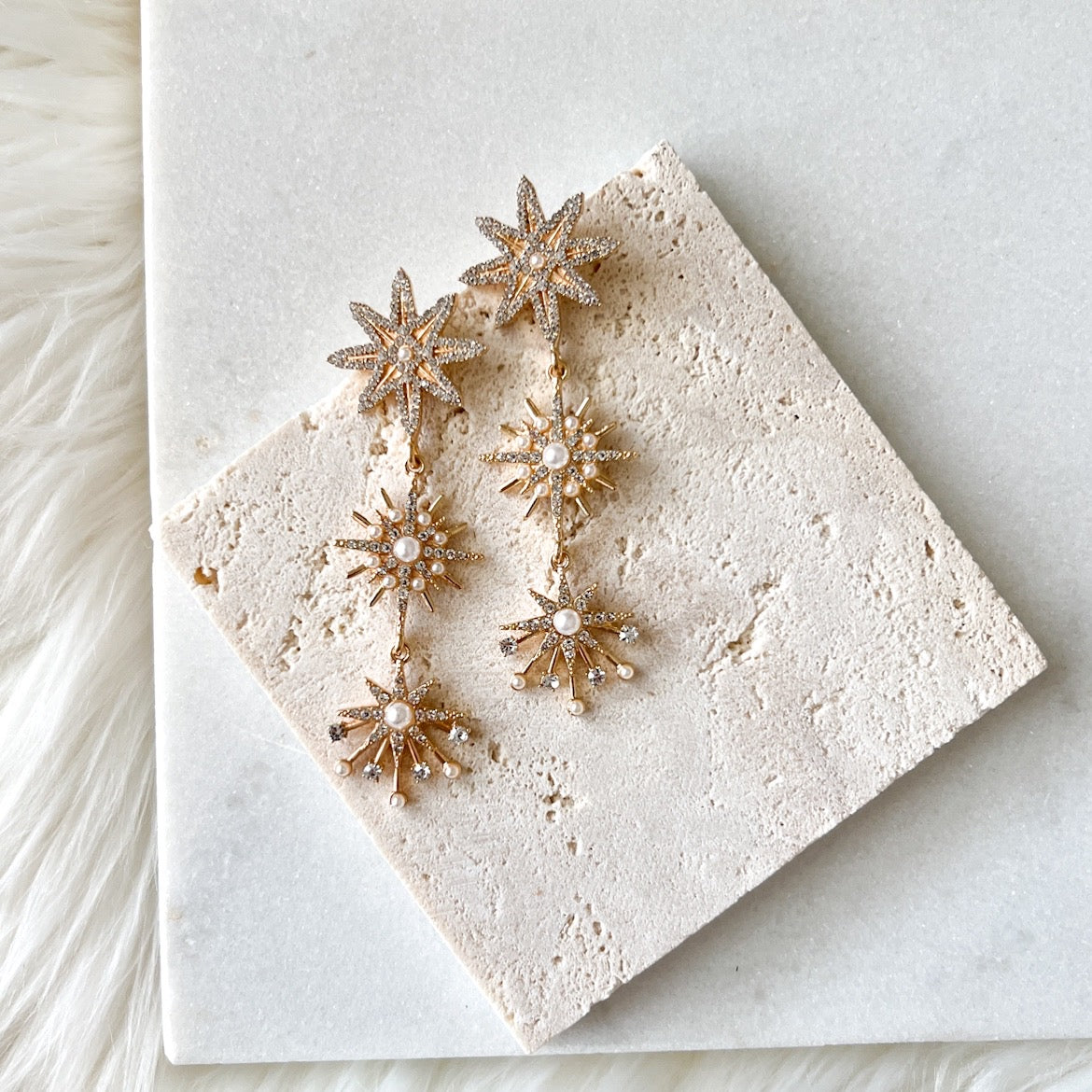 A pair of gold star-shaped dangle earrings with multiple layers, displayed on a white, textured surface.